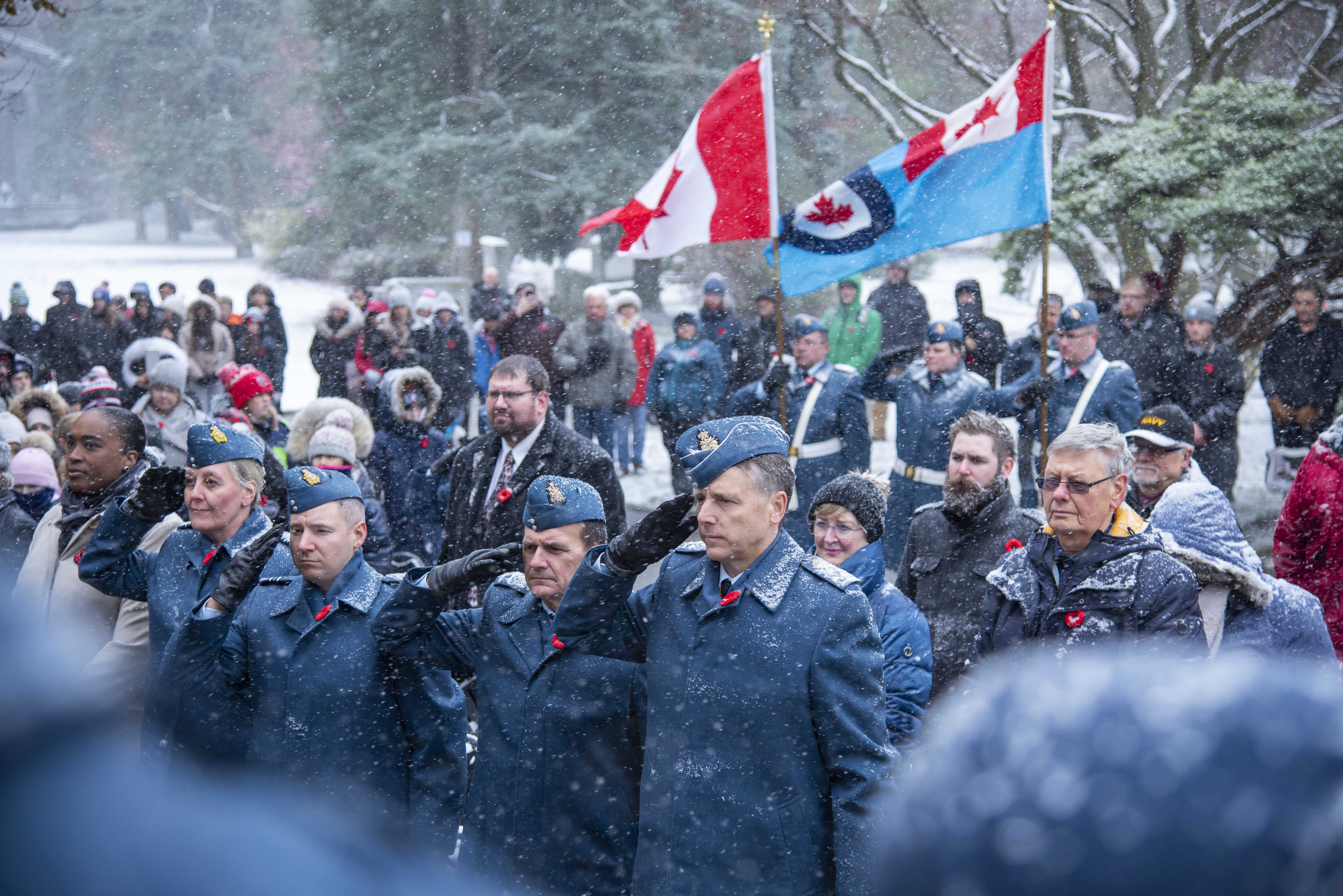 Au milieu d’une foule, quatre personnes vêtues d’uniformes de la Force aérienne saluent, sous la neige qui tombe. 