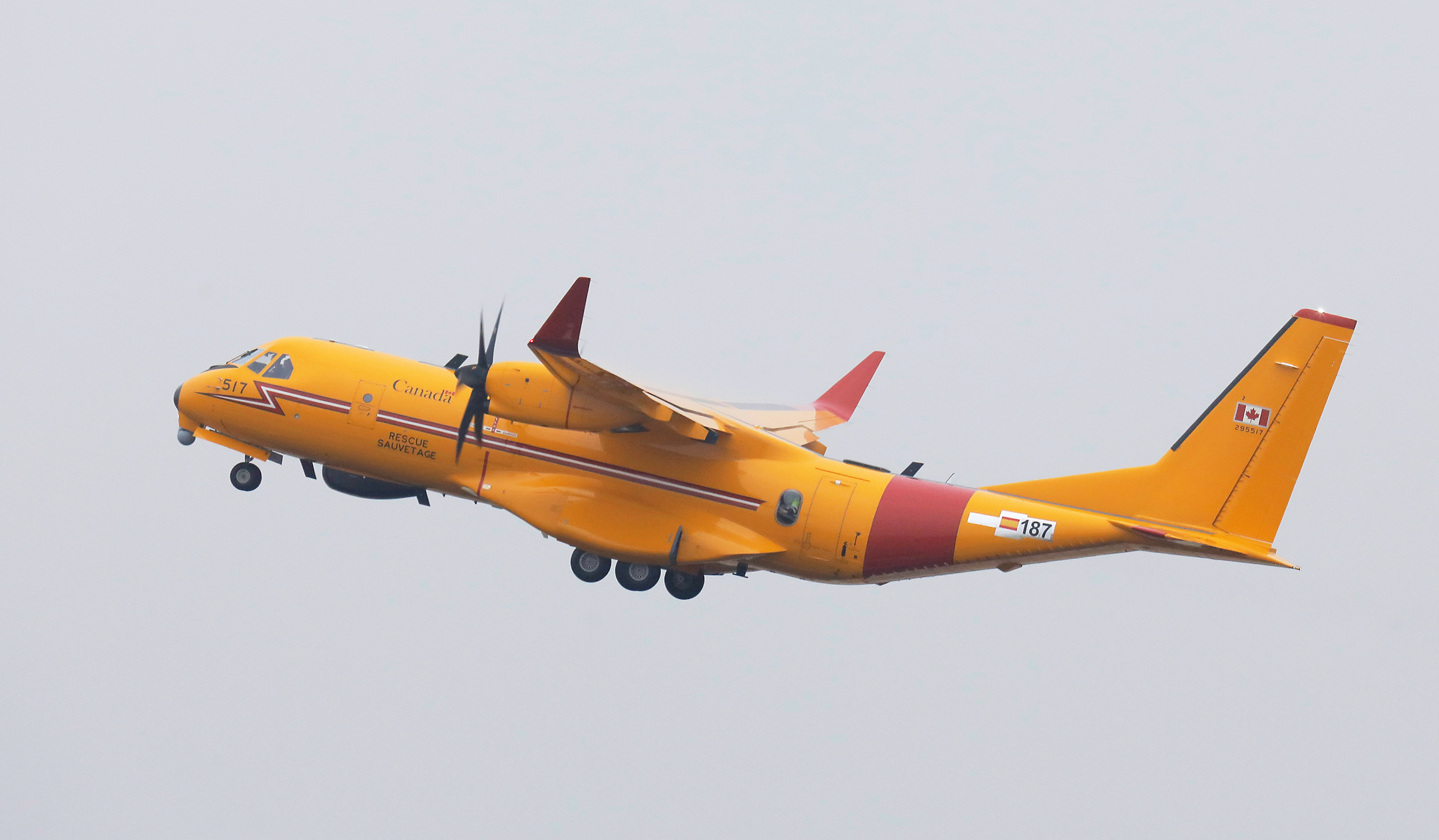 An orange aircraft with red markings ascends in a grey sky.