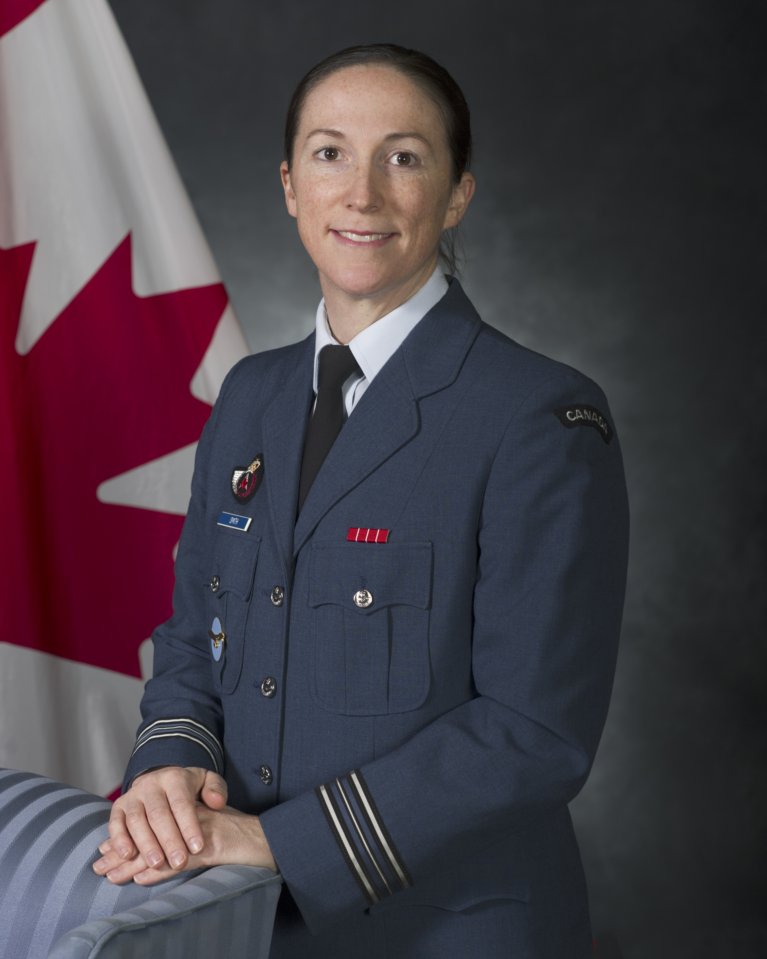 Une femme souriante vêtue d’un uniforme militaire bleu se tient devant un drapeau du Canada, ses mains reposant sur une chaise bleue.
